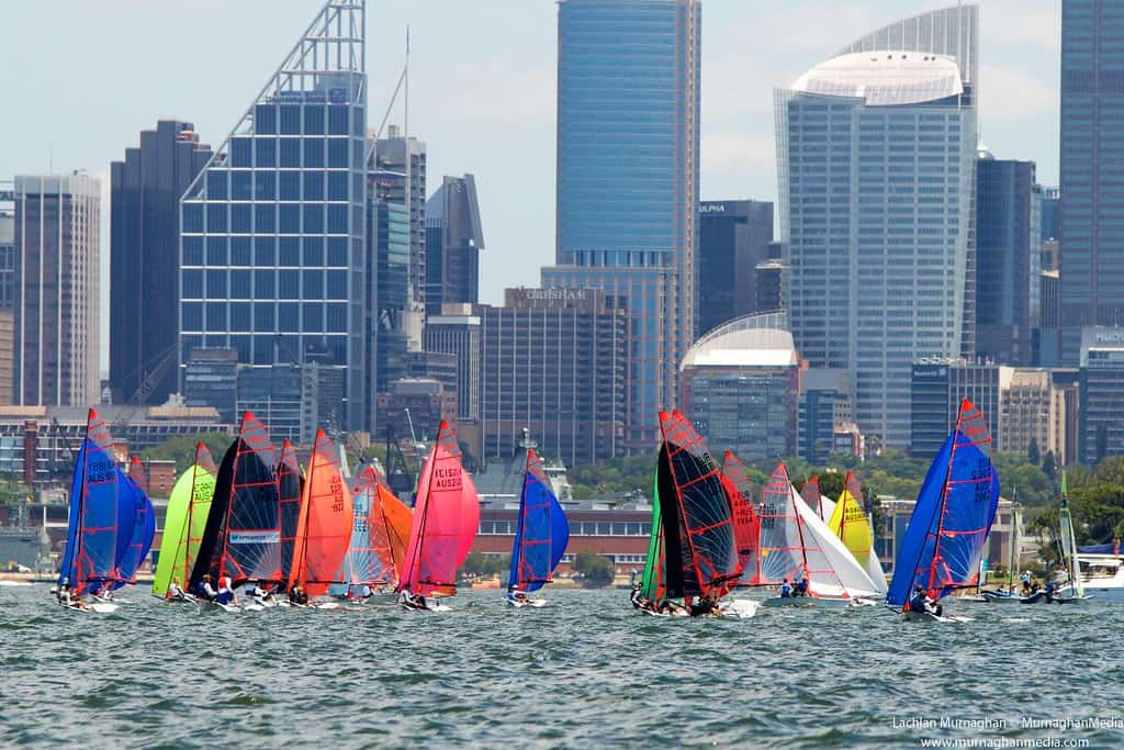 Colorful sailboats racing on Sydney Harbour with city skyline in background.