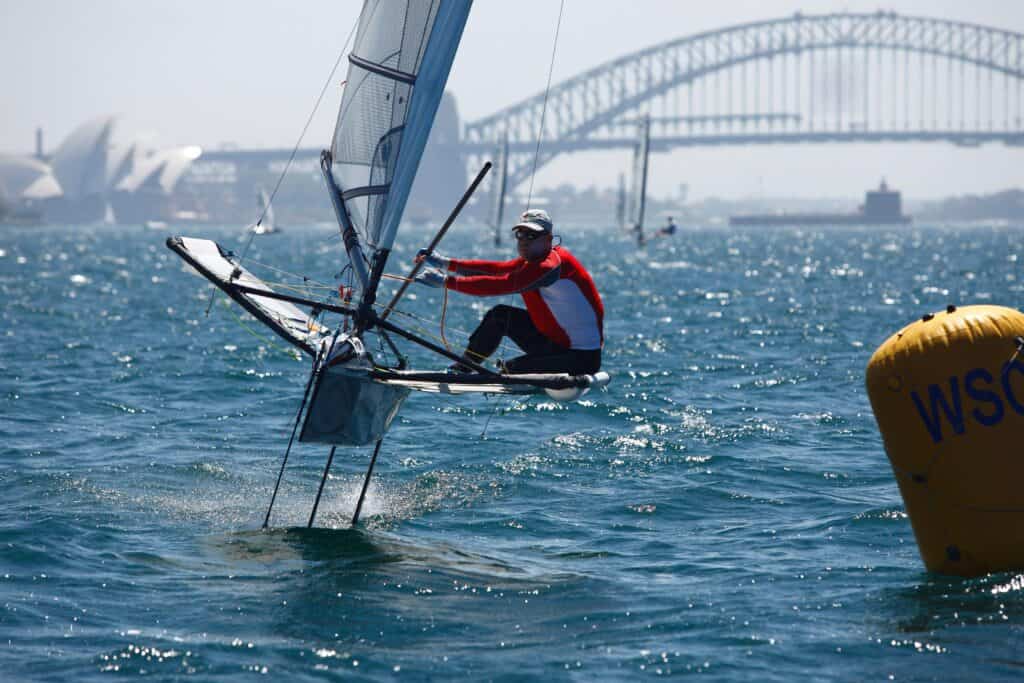 Sailor in red jacket racing a sailboat near Sydney Harbour Bridge.